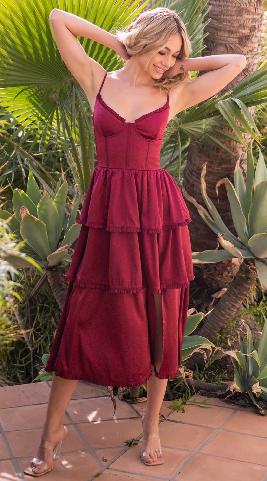 A woman wearing the Velvi Lace-Up Layered Midi Party Dress Maeve stands outdoors on a tiled surface among green plants, lifting her hands to her hair.